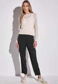 Leichter, cremefarbener Strickpullover mit offenem Muster, kombiniert mit schwarzen Cargo-Hosen mit Seitentaschen und einer lockeren Passform. Weiße Sneakers rundet den Look ab.
