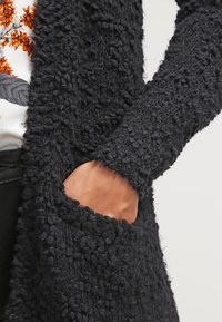 Cardigan noir en texture avec une surface douce et bouclée, doté de poches latérales, avec des manches ajustées et un design à devant ouvert.