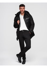 Hombre vistiendo una chaqueta acolchada negra, una camiseta polo blanca, pantalones negros y zapatos negros, de pie con una pierna cruzada en un entorno de estudio.