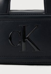 Černá kožená kabelka s lesklým logem "cK". Má texturovaný povrch, čisté linie a kontrastní matné lemování po okrajích.