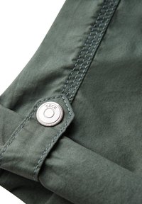 Groene stof met een gladde textuur, voorzien van een zilveren metallic knoop en contrasterende blauwe stiksels langs de rand.