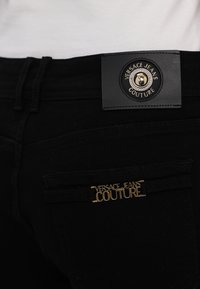 Black denim jeans featuring a large black leather waistband label with a gold emblem and a smaller gold logo accent on the back pocket.