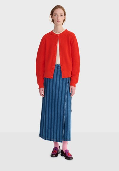 Femme portant un cardigan rouge vif boutonné, une longue jupe en denim à rayures bleues, des chaussettes roses et des mocassins à glands violet foncé, debout devant un fond uni.