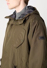 Casaco parka verde-azeitona com forro xadrez cinza, apresentando dois bolsos frontais, capuz com cordão e um emblema no manga.