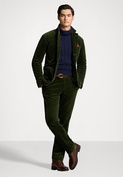 Grønn kordfløyelsdrakt med en tettsittende jakke og matchende bukser, kombinert med en teksturert marineblå genser, brune lærsko og belte.