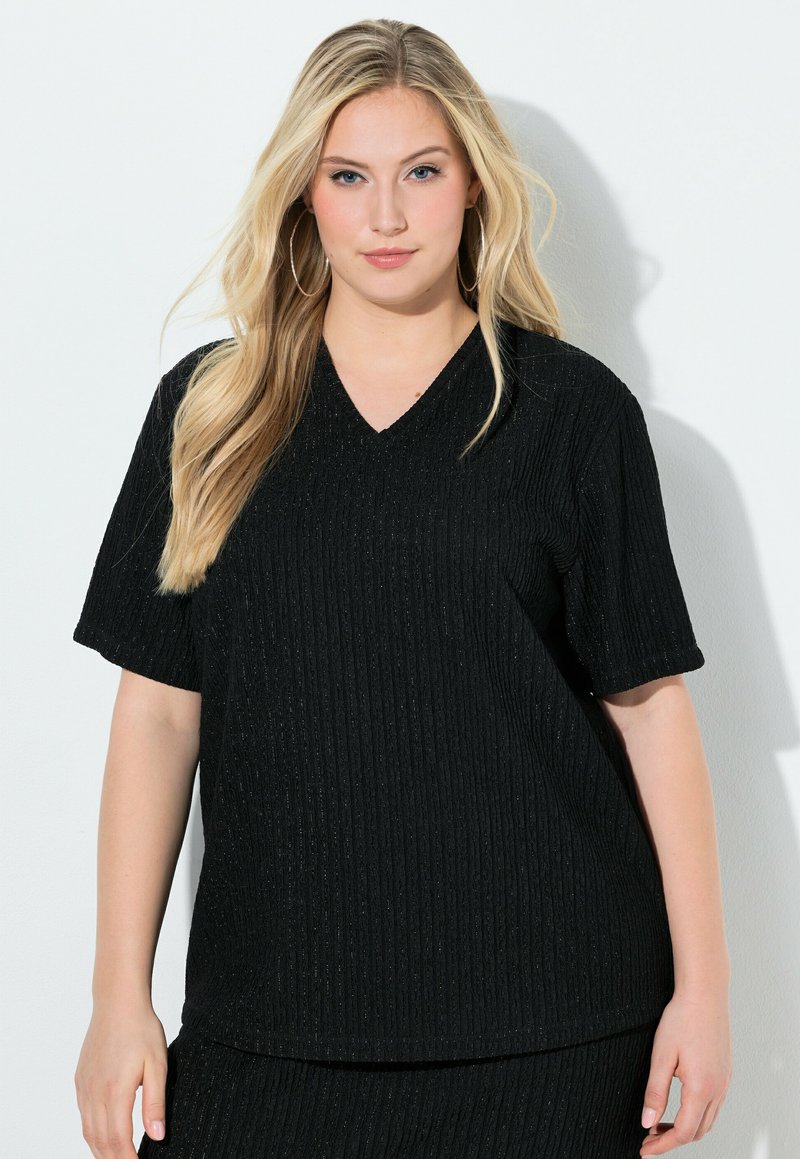 Blondynka o długich włosach, nosząca kolczyki-koła oraz czarną, krótkorękawową bluzkę z teksturą i dekoltem w serek, stojąca na białym tle.