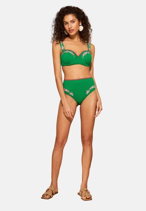 Femme aux cheveux bouclés portant un haut de bikini vert brodé de fleurs et un bas taille haute, des sandales dorées et des boucles d'oreilles blanches en forme de fleur, debout.
