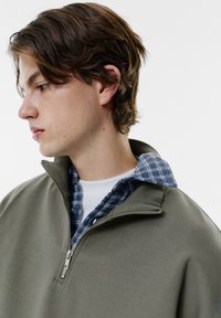 Felpa verde oliva con zip e colletto alto, indossata sopra una camicia a quadri blu. Texture liscia con dettagli in metallo della zip.