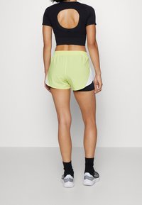 Kvinna som står vänd bort, iklädd svart crop-top med cirkulär ryggskärning, neongula träningsshorts, svarta strumpor och silverfärgade sneakers.