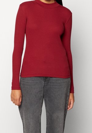 Woman wearing a fitted, long-sleeve, ribbed red sweater paired with high-waisted faded black jeans against a plain background.