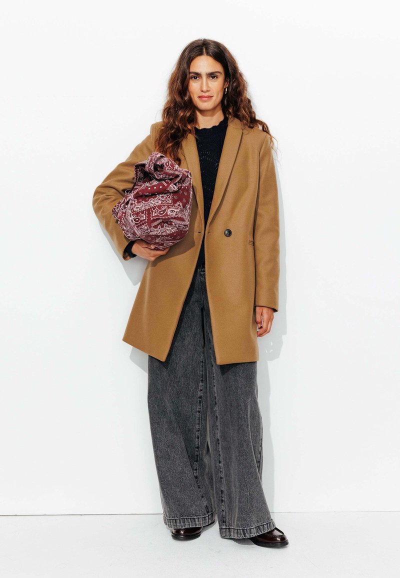 Manteau en laine marron sur un pull noir texturé, associé à un pantalon en denim gris ample. Tient un sac à motif bandana bordeaux.