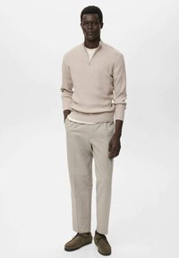 Suéter de punto beige con cremallera sobre una camisa blanca, combinado con pantalones beige claros y zapatos verde oliva, de pie sobre un fondo neutro.