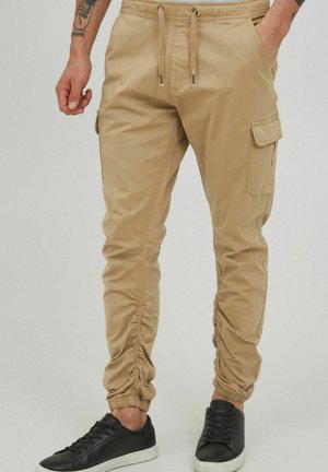 Pantalones cargo beige con puños elásticos, cintura con cordón, bolsillos laterales y detalles fruncidos a los lados. Combinados con zapatillas negras.