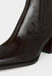 Bottine en cuir brun foncé brillant avec bout pointu, coutures visibles, panneau latéral élastique et petit talon bloc sur fond blanc.