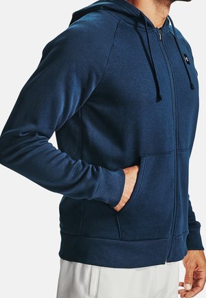 Mann trägt einen marineblauen Hoodie mit Reißverschluss und Kordelzug, hat eine Hand in der Tasche und kombiniert ihn mit hellen Hosen.