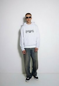 Szara bluza z kapturem z graficznym napisem, zestawiona z luźnymi dżinsami. Biały pasek i czarne sneakersy dopełniają strój.