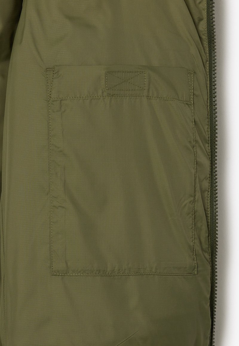 Interior de chaqueta de nailon verde con un bolsillo cuadrado, cierre de cremallera y costuras planas, que destaca una textura suave y ligera.