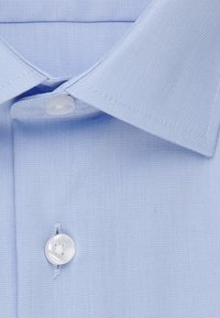 Gros plan sur le col d'une chemise bleu clair avec le bouton supérieur fermé et la texture du tissu visible.