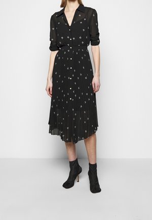 Kobieta w czarnej, sięgającej do połowy długości, zapinanej na guziki sukience z małym białym kwiatowym wzorem, do tego czarne botki na obcasie.