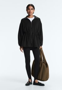 Ženska, oblečena v črno suknjič z zadrgo in črnimi črtami, črne legice in črne čevlje, drži veliko olivno zeleno torbo na belem ozadju.