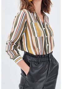 Chemise à rayures vertes, jaunes et blanches avec deux poches poitrine et des manches longues. Pantalon taille haute en cuir noir avec taille élastique.