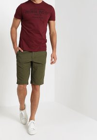T-shirt maroon à col rond avec impression noire, assortie à un short vert. La tenue est complétée par des baskets blanches.