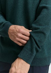 Pulover texturat verde cu mâneci lungi; prim-plan al unei mâini care ajustează manșeta, arătând un detaliu modelat la încheietură.