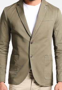 Marynarka w kolorze oliwkowej zieleni wykonana z lekkiego materiału, wyposażona w dwie przednie kieszenie, wycięte klapy oraz jedno zapięcie na guzik. Mały detal w postaci emblematu kotwicy.