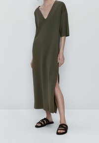 Olive green, knee-length dress with a deep V-neck, short sleeves, and side slits. Styled with black woven sandals. Smooth fabric texture.