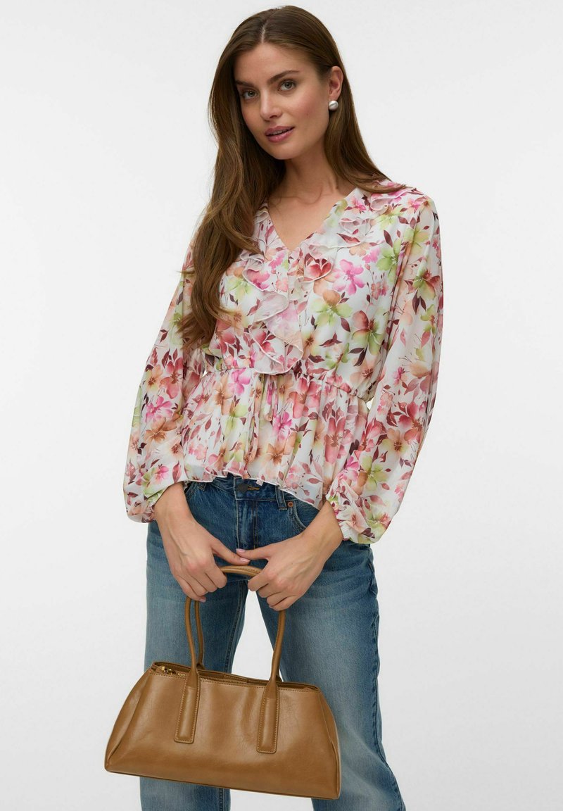 Jeune femme en blouse à volants fleurie et jean, tenant un sac à main en cuir marron sur fond blanc uni.