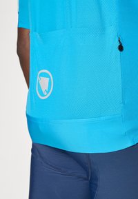 T-shirt de sport turquoise avec un tissu texturé, poche intégrée et un logo circulaire en blanc. Associé à un short de sport bleu marine.