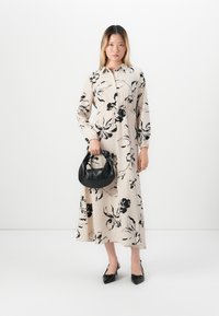 Femme debout dans une longue robe florale beige, tenant un sac à main noir, portant des escarpins noirs à bout pointu, sur fond blanc uni.
