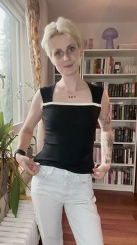Henkilö, jolla on lyhyet vaaleat hiukset ja tatuointeja, säätää mustaa ykköstä ja valkoisia farkkuja huoneessa, jossa on kirjahylly ja ikkuna kasvien kanssa.