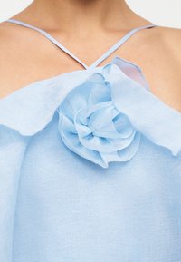 Jasnoniebieski top bez rękawów z cienkimi skrzyżowanymi ramiączkami i dużym materiałowym kwiatem na wysokości klatki piersiowej.