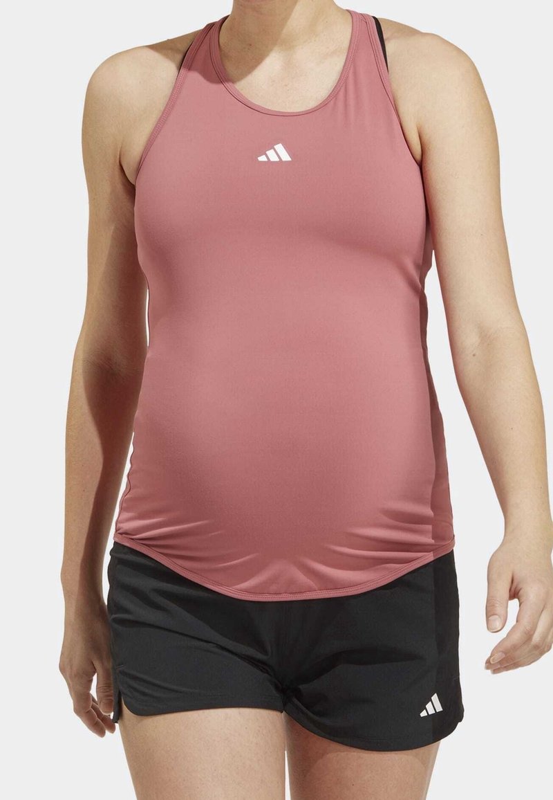Personne portant un haut de sport rose ajusté sans manches et un short noir, debout devant un fond uni.