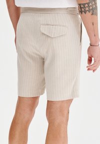 Beżowe spodenki w paski wykonane z lekkiego materiału, z tylną kieszenią z zaokrąglonym klapkiem i prostym dołem. Teksturowane poziome paski.