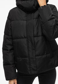 Casaco puffer preto com colarinho alto, design acolchoado e corte solto. O material parece suave e ligeiramente brilhante.