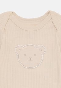 Combinaison beige à côtes avec une appliqué de visage d'ours sur le devant, offrant une texture douce et un col rond.