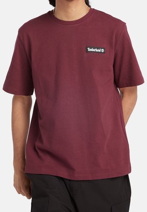 Mand iført en ensfarvet rødbrun Timberland t-shirt med et lille logo på brystet og sorte cargobukser, standing med den ene hånd bag ryggen.