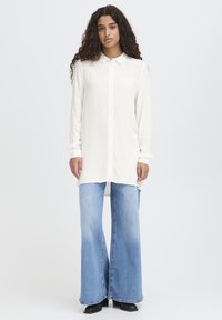 Sieviete ar gariem cirtainiem matiem, tērpusies baltā kreklā ar pogām, gaiši zilās plašām biksēm un melnās augstpapēžu kurpēs, stāv uz baltas fona.