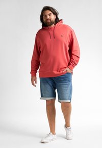 Felpa con cappuccio color corallo con tasca frontale, abbinata a pantaloni shorts in denim e sneakers bianche. La felpa presenta un piccolo emblema vicino al petto.