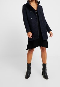 Cappotto blu navy in misto lana con colletto strutturato, design doppiopetto e grandi bottoni neri, abbinato a un vestito nero e stivali con tacco.