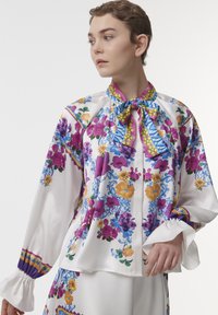 Floral-patterned blouse in smooth, white fabric. Features purple, yellow, and blue designs with a bow collar and ruffled cuffs.