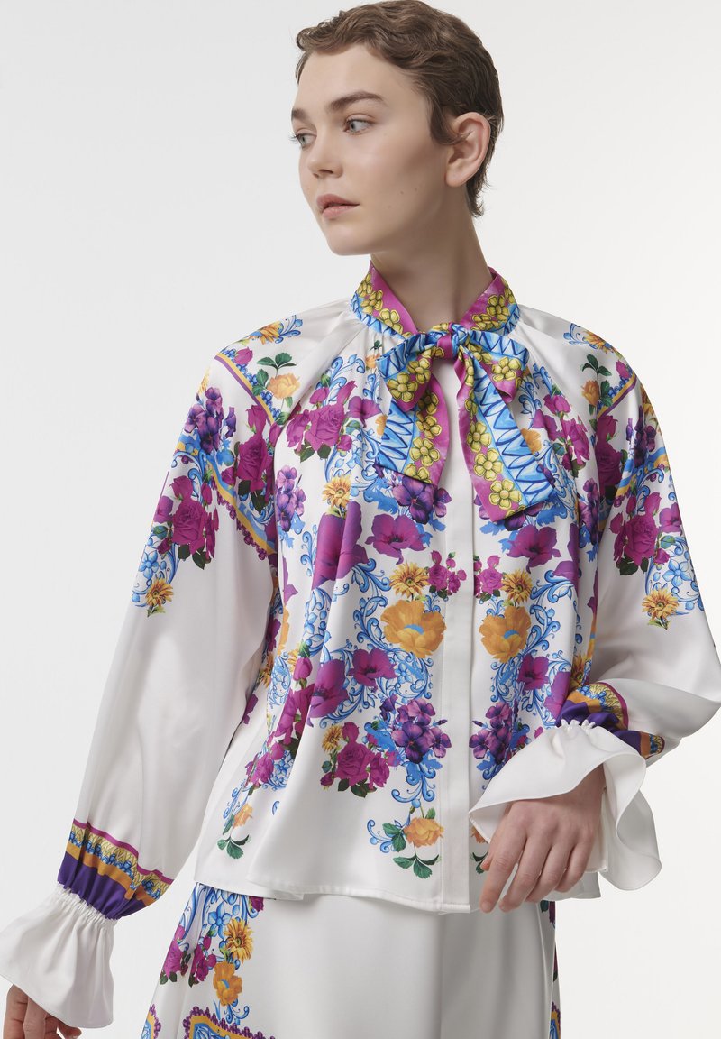 Floral-patterned blouse in smooth, white fabric. Features purple, yellow, and blue designs with a bow collar and ruffled cuffs.