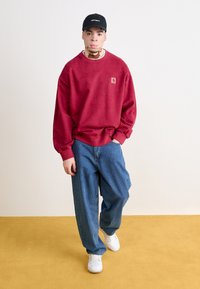 Felpa rossa con una patch logo, indossata sopra una camicia bianca, abbinata a jeans larghi blu e sneakers bianche. Cappellino nero indossato di lato.