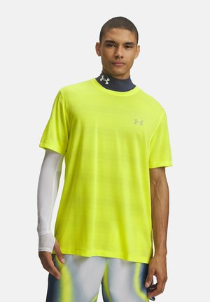 Man die een neongeel mesh Under Armour T-shirt, blauwe nekwarmer, witte mouw en blauw-gele sportshorts draagt, staand tegen een effen achtergrond.