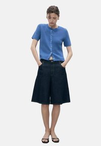 Cardigan en maille côtelée bleue à manches courtes, avec un col rond et une fermeture à boutons à l'avant. Associé à des culottes en denim foncé et des sandales noires.