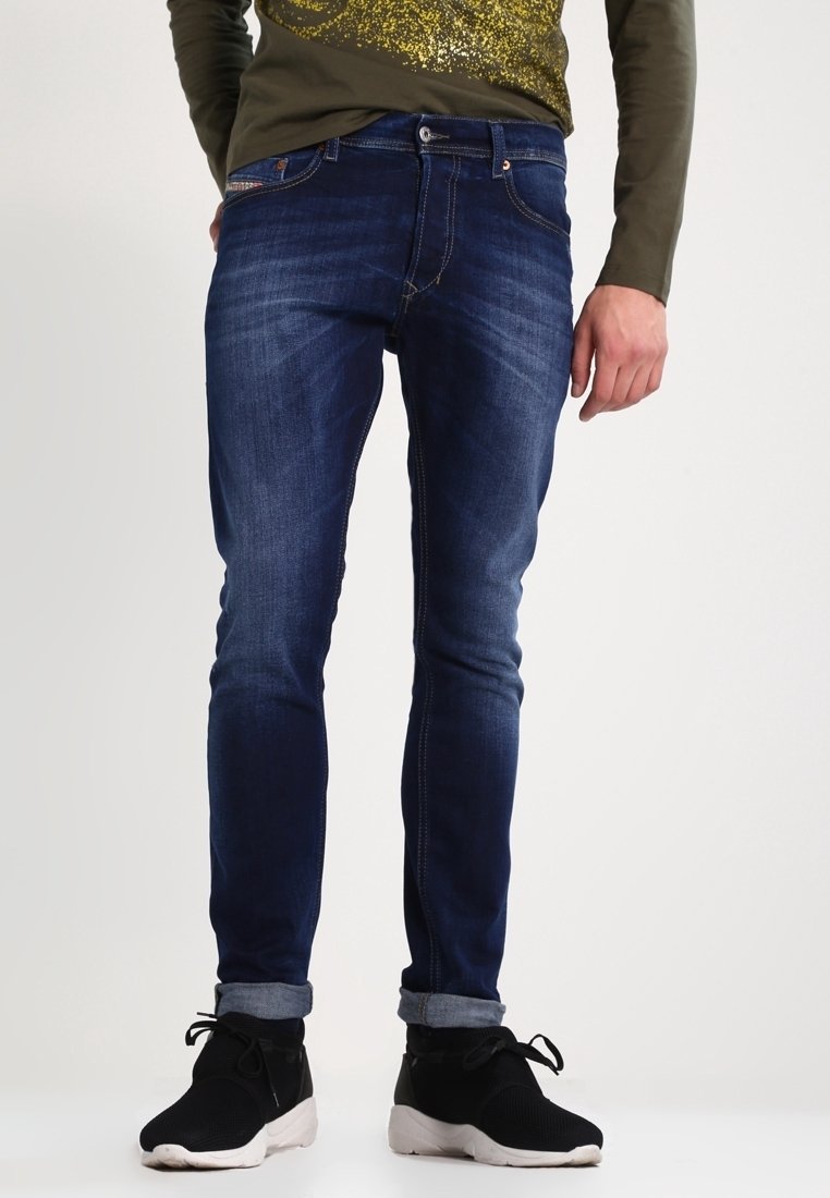 Mörkblå denimjeans för män med slim fit, subtila blekningspartier, sydda detaljer och upprullade avslut. Parat med svarta sneakers.