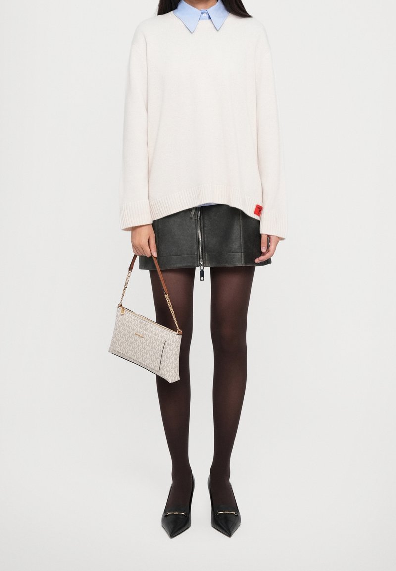 Femme portant un pull blanc par-dessus une chemise bleue à col, une mini-jupe en cuir noire, des collants noirs, des talons pointus noirs, et tenant un sac à main beige à motif.