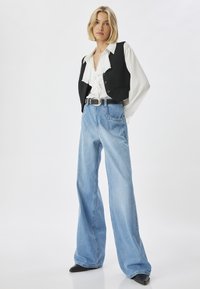 Chaleco negro sobre una blusa blanca con volantes, combinado con unos pantalones de mezclilla de pierna ancha de color azul claro y un cinturón negro. Botas negras de punta completan el look.